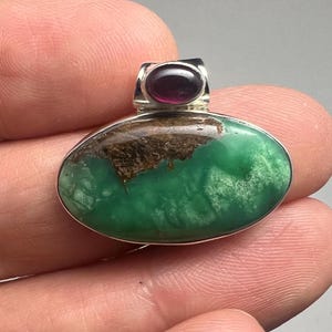 May include: Oval pendant with a vibrant green stone, accented with brown inclusions and a small, dark red stone at the top. The pendant has a silver-colored metal setting.