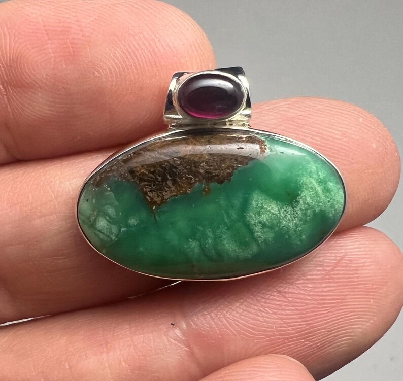 May include: Oval pendant with a vibrant green stone, accented with brown inclusions and a small, dark red stone at the top. The pendant has a silver-colored metal setting.