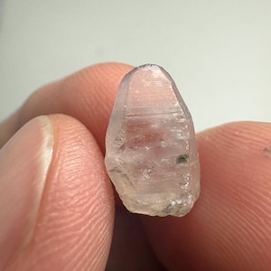May include: A small, translucent, pale lavender-coloured gemstone with a tapered, elongated shape. The surface of the stone displays visible horizontal striations and natural facets, with a subtle, irregular texture and a light, clear appearance.