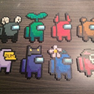 May include: Nine different colored Among Us characters made with perler beads. The characters are gray, green, red, brown, purple, blue, and pink. Each character has a different accessory, such as a plant, a flower, or a hat.