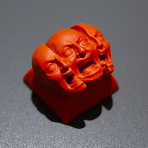 May include: A vibrant orange sculpture featuring three expressive faces. The faces are carved with detailed features and appear to be in a state of distress or emotion. The sculpture is mounted on a square base.