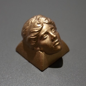 May include: A gold-colored sculpture of a classical female bust. The bust is set on a square base and has detailed facial features and flowing hair. The sculpture is made of a metallic material.