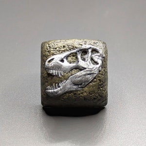 Könnte beinhalten: Ein kleiner, quadratischer, goldfarbener Harzblock mit einem silberfarbenen Dinosaurierschädel, der auf der Vorderseite geprägt ist.