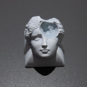 May include: A white sculpture of a classical female head with a portion of the head carved away to reveal a spiral design. The sculpture is set against a gray background, creating a stark contrast.