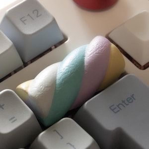 May include: A close-up of a white mechanical keyboard with light gray keycaps. A red cylindrical object sits on the top right. A colorful, twisted marshmallow rests on the keyboard between the keys.