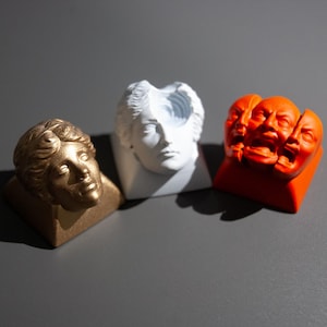 May include: Three decorative busts in gold, white, and orange. Each bust features a classical human face. The gold bust is on the left, the white bust is in the middle, and the orange bust is on the right. The busts are on a gray surface.