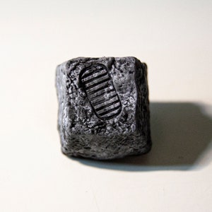 May include: A small, gray, cube-shaped rock with a raised footprint design.