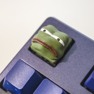 May include: A green frog-shaped keycap with a brown mouth and white eyes, sitting on a blue keyboard.