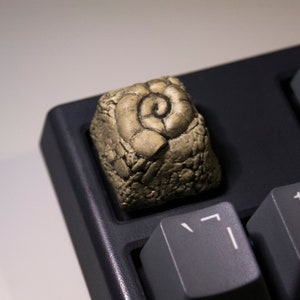 May include: A gold-coloured keycap with a detailed spiral design, resembling a seashell, sits on a black keyboard.
