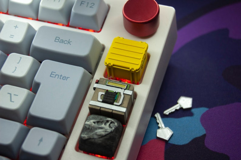 May include: A white mechanical keyboard with custom keycaps. The keycaps are themed around a military crate, a dinosaur, and a key. The keyboard is on a purple and blue background.