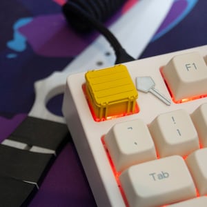 May include: A white mechanical keyboard with a yellow keycap shaped like a shipping container. The keyboard has red backlighting and the keycaps are white with black lettering. The keycap is on the '7' key.