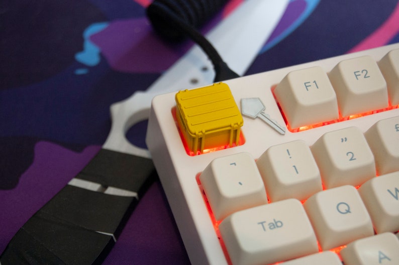 May include: A white mechanical keyboard with a yellow keycap shaped like a shipping container. The keyboard has red backlighting and the keycaps are white with black lettering. The keycap is on the '7' key.