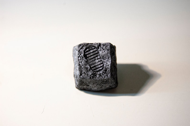 May include: A small, gray, cube-shaped rock with a raised footprint design.