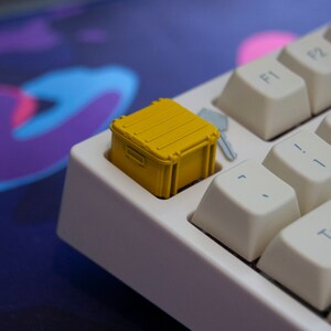 May include: A yellow plastic container keycap for a mechanical keyboard. The keycap is shaped like a small box with a lid and is sitting on a white keyboard.
