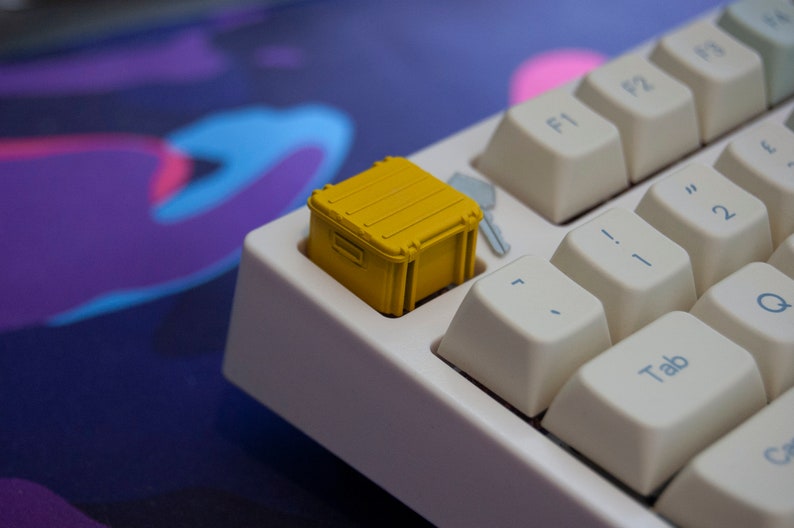 May include: A yellow plastic container keycap for a mechanical keyboard. The keycap is shaped like a small box with a lid and is sitting on a white keyboard.
