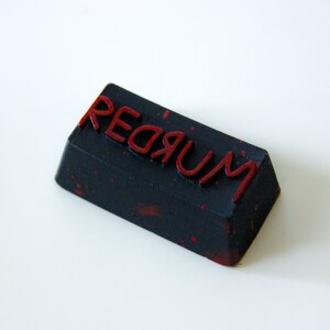May include: A black rectangular keycap with the word "REDRUM" written in red lettering. The keycap has a speckled, textured surface.