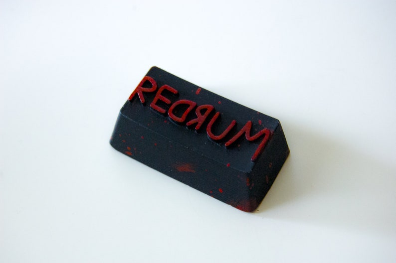 May include: A black rectangular keycap with the word "REDRUM" written in red lettering. The keycap has a speckled, textured surface.