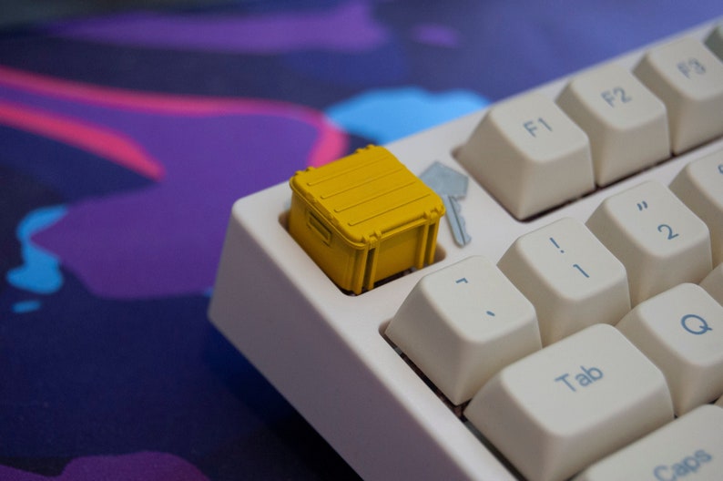 May include: A yellow keycap shaped like a small, rectangular container with a latch. The keycap is on a white keyboard with keys labelled 'Tab', 'Q', '2', '!', 'F1', and 'F2'.