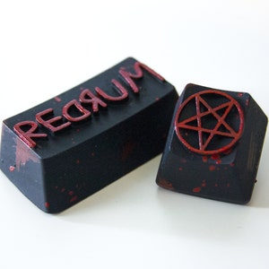May include: Two black keycaps with red paint splatters. One keycap has the word "REDRUM" printed in red letters. The other keycap has a red pentagram.