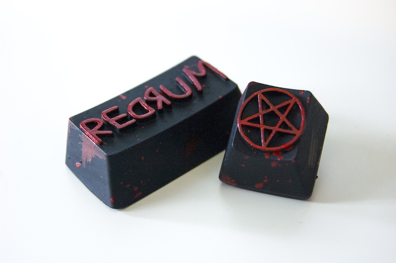May include: Two black keycaps with red paint splatters. One keycap has the word "REDRUM" printed in red letters. The other keycap has a red pentagram.