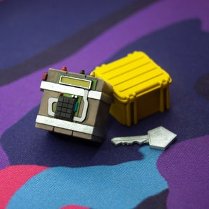 May include: A small, brown, box-shaped device with a digital display and buttons, a yellow plastic case, and a silver key. The items are on a purple and blue patterned surface.