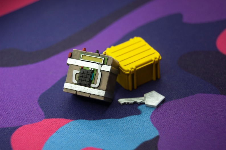May include: A small, brown, box-shaped device with a digital display and buttons, a yellow plastic case, and a silver key. The items are on a purple and blue patterned surface.