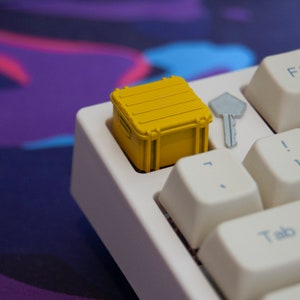 May include: A yellow keycap shaped like a small shipping container sits on a white keyboard. The keycap is on the key to the left of the '1' key.