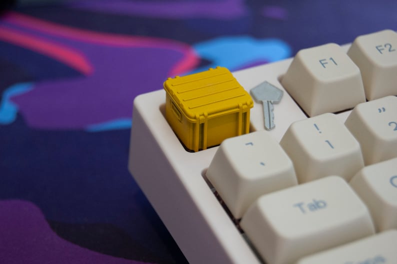 May include: A yellow keycap shaped like a small shipping container sits on a white keyboard. The keycap is on the key to the left of the '1' key.