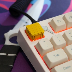 May include: A white mechanical keyboard with a yellow container keycap. The keyboard has a red backlight and the keys are labelled with letters and numbers. The container keycap is a small, yellow plastic container with a lid.