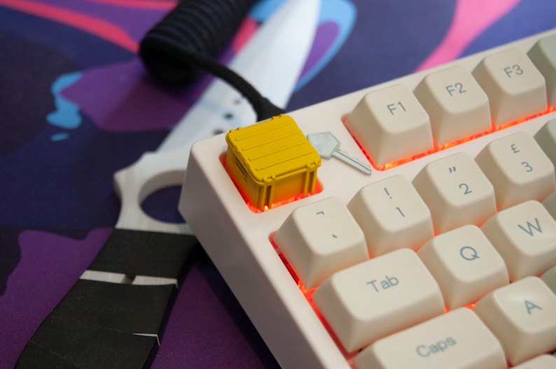May include: A white mechanical keyboard with a yellow container keycap. The keyboard has a red backlight and the keys are labelled with letters and numbers. The container keycap is a small, yellow plastic container with a lid.
