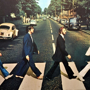 Puede incluir: Una fotografía en blanco y negro de cuatro hombres caminando por un paso de cebra. Los hombres visten trajes y pantalones vaqueros. La foto está tomada desde un ángulo bajo, y los hombres caminan hacia la cámara. La foto es una famosa portada de álbum de los Beatles.
