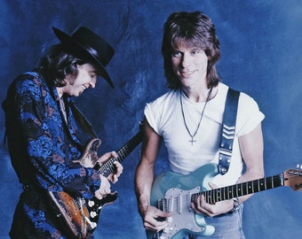 May include: Two men with guitars pose for a photo. One wears a black hat and a patterned jacket, the other a white t-shirt. Both hold electric guitars, one a light blue Fender Stratocaster.