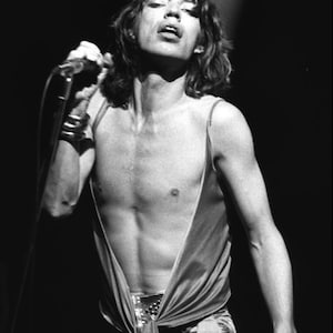 Puede incluir: Imagen en blanco y negro de una persona actuando en el escenario, sosteniendo un micrófono. La persona lleva un atuendo de satén con un escote pronunciado y un cinturón. La persona tiene el pelo largo y lleva una pulsera en el brazo izquierdo.