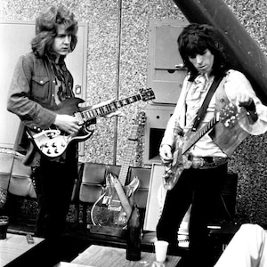 May include: Two musicians, one with long hair, playing electric guitars in a black and white photograph. The musician on the left is wearing a denim jacket and the musician on the right is wearing a white shirt with a vest.