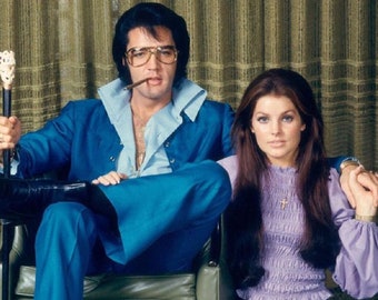 May include: A vintage photograph of a man in a blue suit, wearing large glasses and smoking a cigar, seated beside a woman in a purple top. The man holds a cane with a decorative handle. The background has a green curtain.
