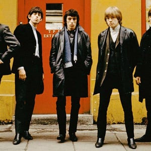 May include: Five men in black suits and coats stand in front of a red door and yellow wall. The men are wearing different styles of suits and coats, and some are wearing scarves. The door has a sign that says "TP ENT".
