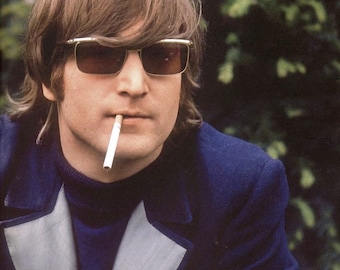 May include: A portrait of a man wearing sunglasses and a dark blue jacket with a light blue trim. He has brown hair and a cigarette in his mouth. The background is a blurred green.