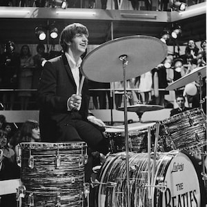 Könnte beinhalten: Ein Schwarzweißfoto von Ringo Starr, dem Schlagzeuger der Beatles, der ein Schlagzeug spielt. Die Bassdrum trägt den Namen der Band, "The Beatles", in weißen Buchstaben. Das Foto wurde während eines Live-Auftritts aufgenommen.