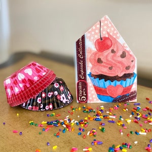 May include: A set of 5 pink and black cupcake liners with a pink and white cupcake with a red cherry on top and the text "5 Cupcakes Cupcake Collection 5 Pcs. Bakery Shop" on a pink and white background.