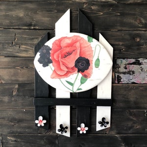 May include: A white and black wooden coat rack with a round, white painted panel featuring a red poppy and black flowers. The panel is surrounded by a black frame. The coat rack has five hooks.