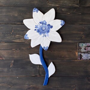 May include: A white ceramic flower with blue and white floral designs. The flower is attached to a blue wooden stem with white ceramic leaves.