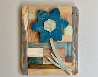 May include: A blue flower with a green stem is made from wood and painted in a patchwork style. The flower is a hexagon shape with six petals. The background is a wooden frame with a patchwork of different colored wood.