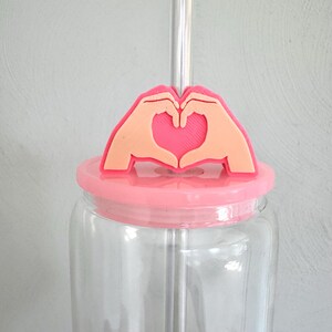 May include: A clear glass jar with a pink lid and a clear straw. The lid features a pink and peach-colored heart-shaped hand gesture design. The jar is empty and sits against a light gray background.