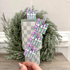 May include: A clear straw with book-shaped charms. The charms are in shades of purple and green, tied with a white bow. The tumbler is checkered in light blue and white. A green wreath is in the background.