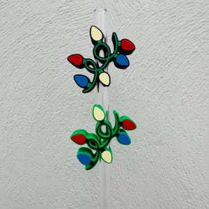 May include: Two Christmas light charms with red, blue, and yellow bulbs on a green vine. The charms are attached to a clear straw, set against a textured white background. Festive holiday decor.