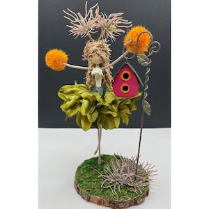 May include: A whimsical fairy figurine with green and brown fabric wings, holding a small red birdhouse with two holes. The fairy is standing on a moss-covered base with a metal stem and orange flower accents.
