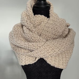 Pode incluir: Cachecol gola bege, feito à mão em crochê, exposto em um manequim preto. O cachecol é feito de um fio grosso, com um padrão de ponto texturizado. A gola é enrolada ao redor do pescoço do manequim, criando um visual quente e elegante.