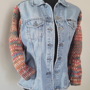 Op de afbeelding: Lichtblauwe spijkerjas met gehaakte mouwen in tinten rood, oranje, blauw en bruin. De jas heeft een klassieke kraag, knoopsluiting en twee borstzakken. Een uniek, handgemaakt kledingstuk.