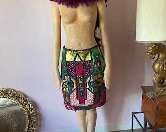 May include: A colourful sequined mini skirt with geometric patterns in shades of pink, green, blue, yellow, and purple. The skirt is worn on a mannequin with a purple feather boa.