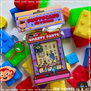 May include: A clear plastic package containing a "Smarty Pants" photocard keychain, featuring a cartoon arcade game design with various character illustrations. The package is surrounded by colorful toy building blocks.